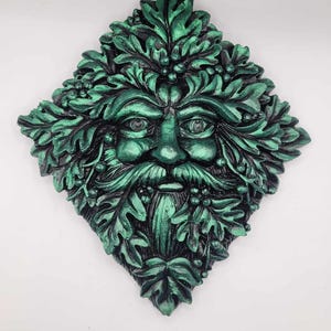 May include: A diamond-shaped wall plaque featuring a green man design. The face, with a beard and surrounded by leaves, is painted in metallic green and black. The plaque is a decorative item.