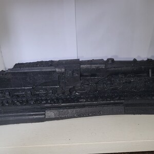 May include: A black model train locomotive on a black base. The train is on a track and has a detailed design.