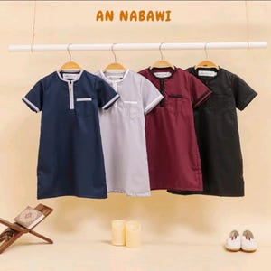 May include: Four short-sleeved tunic shirts in navy blue, gray, burgundy, and black hang on a clothes rack. Each shirt has a mandarin collar, a chest pocket, and a buttoned placket. The text "AN NABAWI" is at the top.
