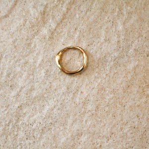 May include: A gold-colored ring with an organic, textured design. The ring is circular and appears to be made of metal. The ring is set against a neutral, textured background, creating a simple and elegant composition.