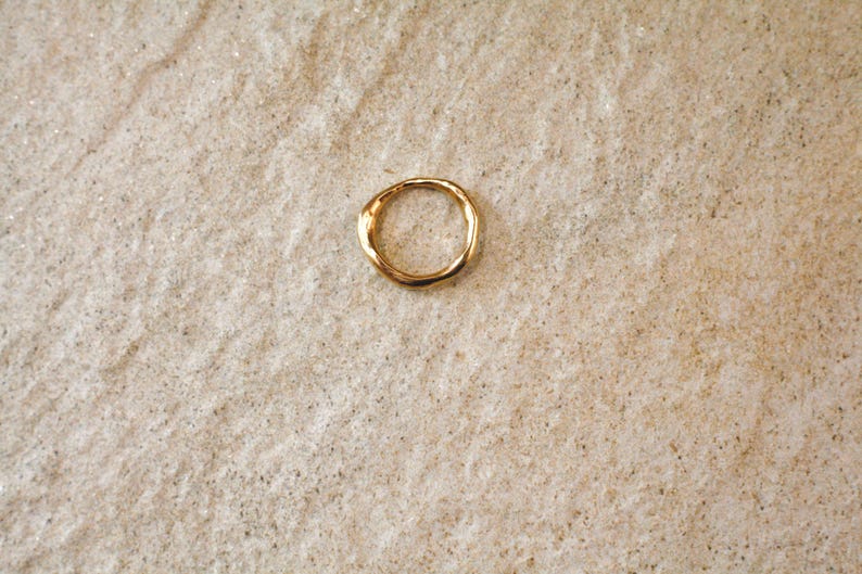 May include: A gold-colored ring with an organic, textured design. The ring is circular and appears to be made of metal. The ring is set against a neutral, textured background, creating a simple and elegant composition.