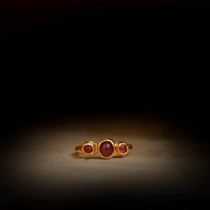 May include: A gold ring with three oval, deep red gemstones. The ring's band is polished gold, and the gemstones are set close together. The ring is displayed on a light surface against a dark background. The gemstones are likely rubies.