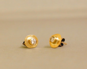 Puede incluir: Un par de pendientes de oro. Cada pendiente presenta una pequeña gema redonda y transparente engastada en un bisel de oro. El oro tiene un aspecto texturizado y martillado. Los pendientes se muestran sobre un fondo neutro.