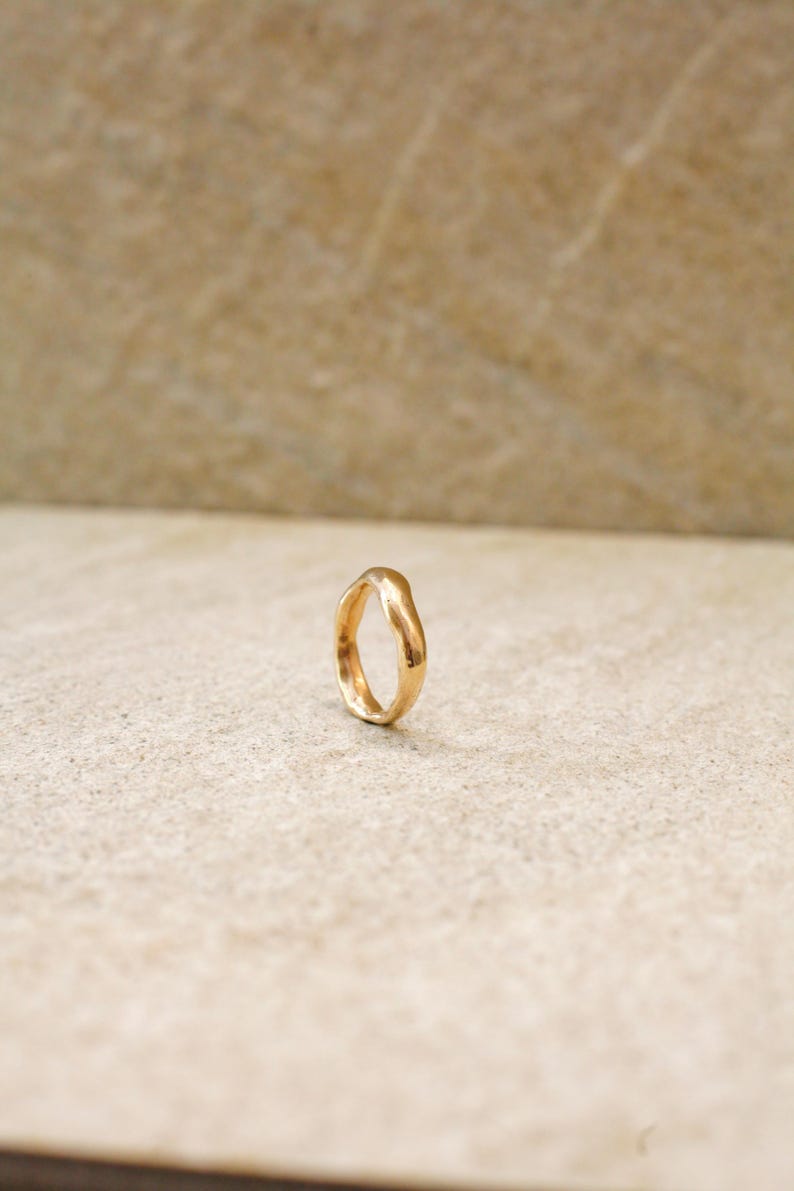 May include: A gold-colored ring with an organic, irregular shape. The ring is set against a neutral background, highlighting its unique design. The ring's surface reflects light, showcasing its polished finish. A piece of jewelry.