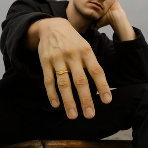 May include: A gold ring with a unique, organic shape. The ring is worn on a finger, and the focus is on the jewelry. The ring has a smooth, polished surface and a slightly irregular design, suggesting a handcrafted aesthetic. The background is blurred.