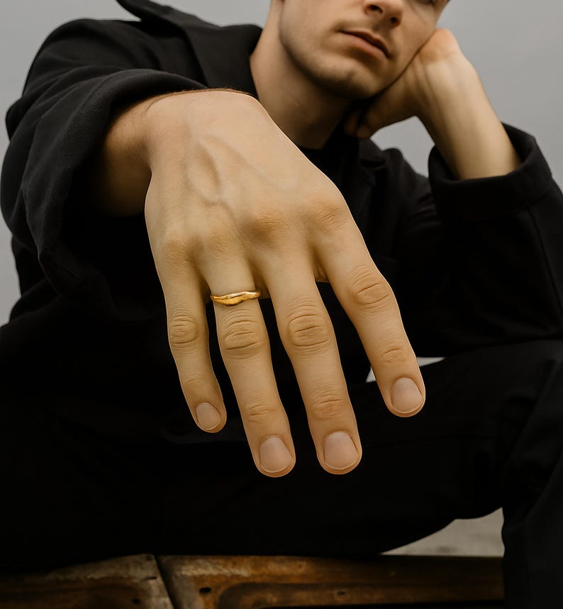 May include: A gold ring with a unique, organic shape. The ring is worn on a finger, and the focus is on the jewelry. The ring has a smooth, polished surface and a slightly irregular design, suggesting a handcrafted aesthetic. The background is blurred.
