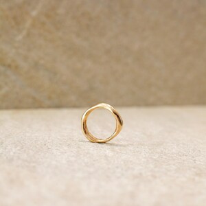 May include: A gold-colored ring with an organic, irregular shape. The ring is set against a neutral, light-colored background. The ring's design features a smooth, polished finish, reflecting the light.