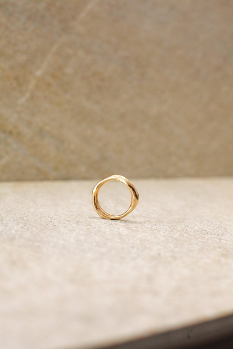 May include: A gold-colored ring with an organic, irregular shape. The ring is set against a neutral, light-colored background. The ring's design features a smooth, polished finish, reflecting the light.