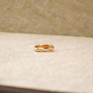 May include: A gold-colored ring with a wavy design. The ring is set against a neutral background, highlighting its unique shape and metallic sheen. The ring's design is simple yet elegant.