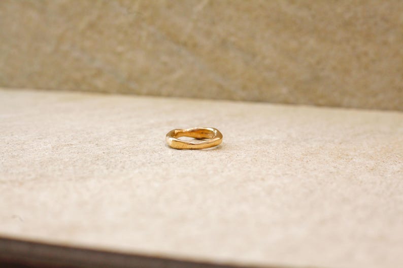 May include: A gold-colored ring with a wavy design. The ring is set against a neutral background, highlighting its unique shape and metallic sheen. The ring's design is simple yet elegant.