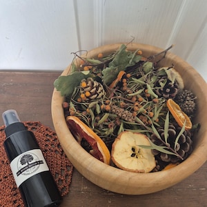 May include: A black spray bottle with a white label and a tree logo, beside a wooden bowl filled with dried fruit, pine cones, and greenery. The bowl is on a brown surface, with a decorative brown doily nearby.