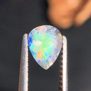Op de afbeelding: Een peervormige, iriserende edelsteen met een regenboog van kleuren, waaronder blauw, groen en geel, vastgehouden in zilveren pincetten.
