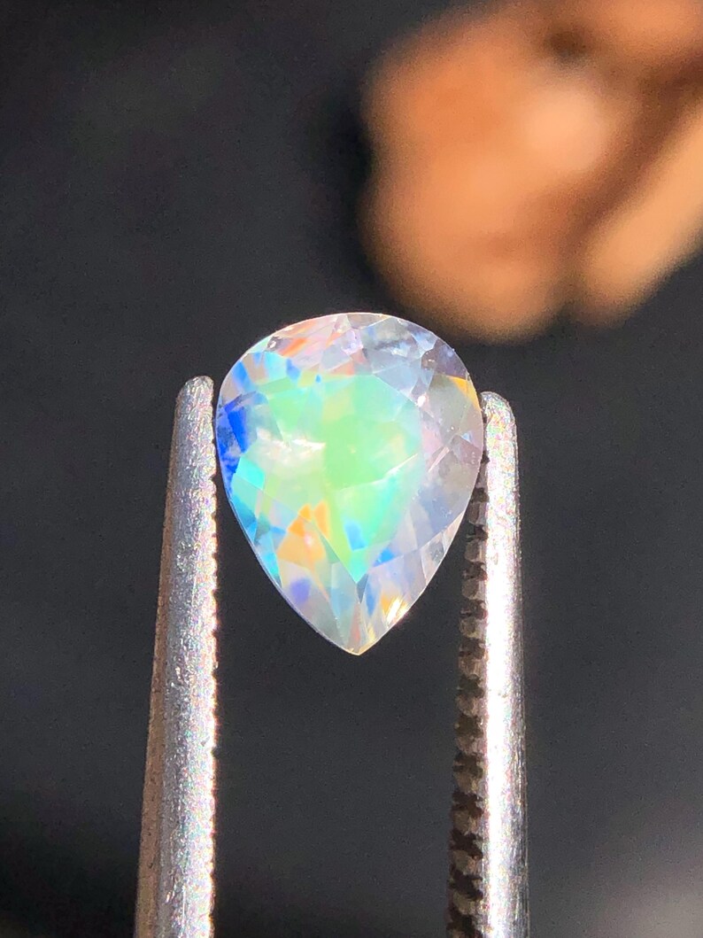 Op de afbeelding: Een peervormige, iriserende edelsteen met een regenboog van kleuren, waaronder blauw, groen en geel, vastgehouden in zilveren pincetten.
