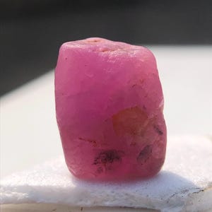 May include: A rough, uncut ruby gemstone. The ruby is a vibrant pink color with a slightly irregular shape. The surface has a natural, textured appearance, with some darker inclusions visible. The gemstone is displayed on a white surface.