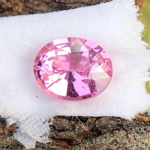 May include: A pink oval-shaped gemstone with a faceted cut. The gemstone is set on a white fabric background.