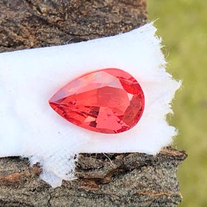 May include: A pear-shaped, red gemstone with a faceted cut. The gemstone is displayed on a white fabric background.