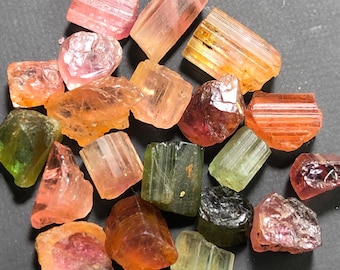 May include: A collection of raw tourmaline gemstones in various colors, including pink, orange, green, and brown. The gemstones have a rough, natural texture and are irregularly shaped. The image is taken from a top-down perspective.