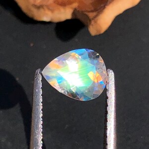 Op de afbeelding: Een peervormige, iriserende edelsteen met een regenboog aan kleuren, vastgehouden in een pincet.