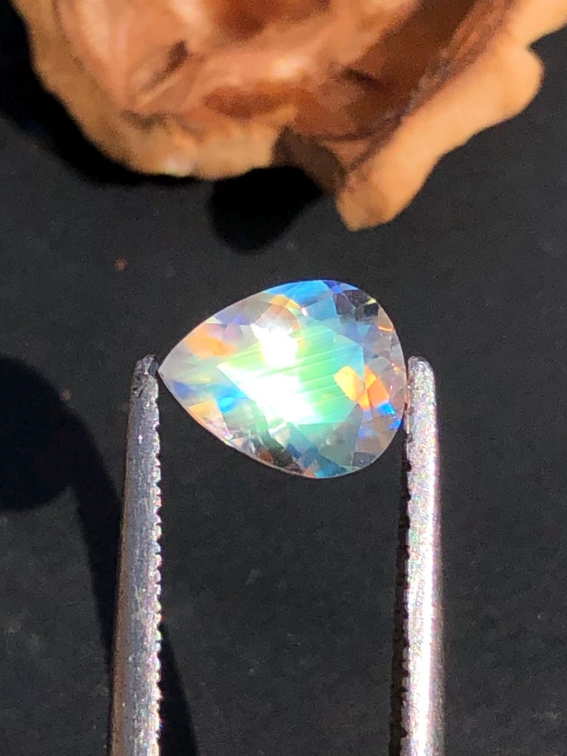 Op de afbeelding: Een peervormige, iriserende edelsteen met een regenboog aan kleuren, vastgehouden in een pincet.