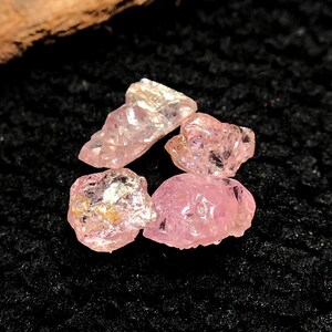May include: Four rough-cut, light pink gemstones are arranged on a black, textured surface. The stones have a crystalline appearance, with varying shapes and sizes. The lighting highlights the facets and internal structures of the gems, creating a sparkling effect.