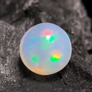 Könnte beinhalten: Ein runder, schillernder Opal-Edelstein mit milchig-weißer Grundfarbe. Der Opal zeigt Farbblitze in Rot, Grün und Gelb. Der Edelstein ist vor einem dunklen, strukturierten Hintergrund platziert, der seine lebendigen Farben hervorhebt.