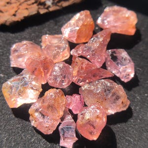 May include: A collection of raw, uncut gemstones in shades of pink and peach. The rough-cut stones vary in size and shape, with a sparkling, translucent quality. The gemstones are scattered on a dark surface, with a piece of wood in the background.