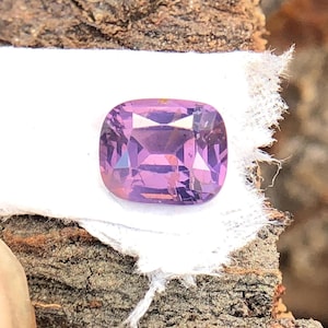 May include: A cushion-cut purple gemstone with facets. The gemstone is on a white surface.
