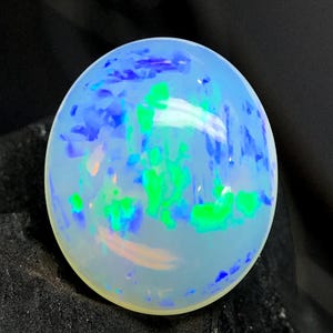 May include: A spherical opal gemstone, exhibiting a milky white hue with internal flashes of blue and green. The polished surface reflects light, showcasing the gemstone's play-of-colour. The opal is set against a dark backdrop.