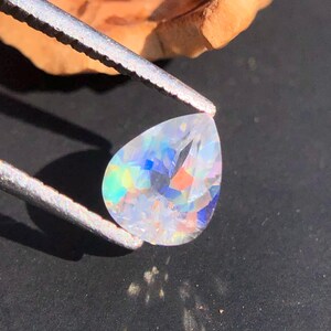 Op de afbeelding: Een peervormige, heldere edelsteen met iriserende regenboogkleuren. De edelsteen wordt vastgehouden door een pincet.