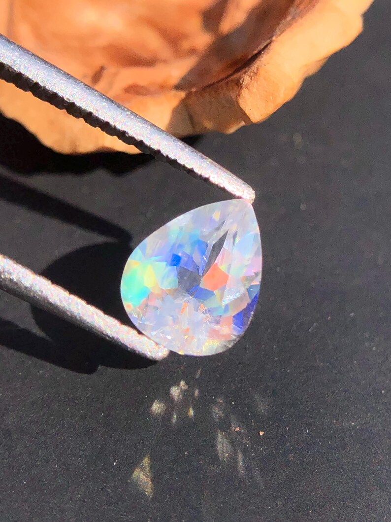 Op de afbeelding: Een peervormige, heldere edelsteen met iriserende regenboogkleuren. De edelsteen wordt vastgehouden door een pincet.