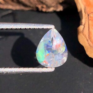 Op de afbeelding: Een peervormige opaal edelsteen met een regenboog aan kleuren, vastgehouden in een pincet tegen een donkere achtergrond.