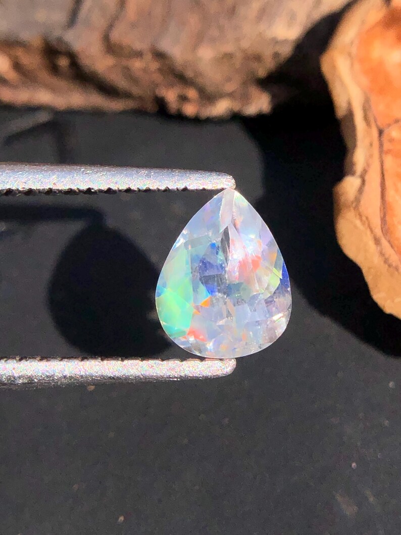 Op de afbeelding: Een peervormige opaal edelsteen met een regenboog aan kleuren, vastgehouden in een pincet tegen een donkere achtergrond.