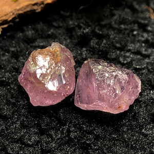 May include: Two rough-cut, raw amethyst crystals. The crystals are a light purple color with a slightly translucent appearance. The crystals are displayed on a black, textured surface. The crystals have a natural, unpolished look.