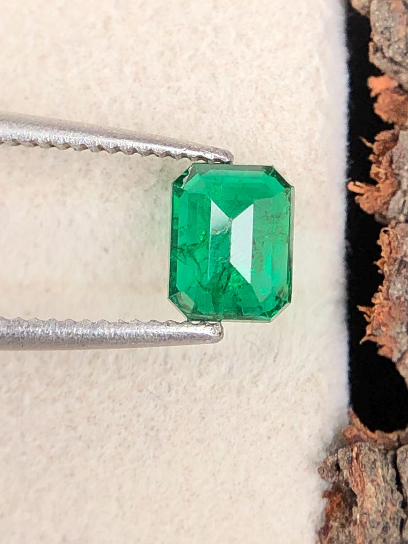 Op de afbeelding: Een levendige, rechthoekige smaragd, vastgehouden door een zilveren pincet. De smaragd heeft een diepgroene tint en een facetgeslepen snede, die het licht reflecteert. De edelsteen wordt gepresenteerd tegen een neutrale achtergrond, wat de helderheid en kleur benadrukt.