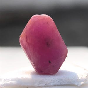 May include: A raw, uncut ruby gemstone. The gem is a vibrant pinkish-red color, with a slightly irregular, faceted shape. It sits on a white surface, with a blurred background.