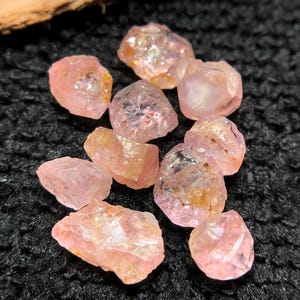 May include: A collection of rough, uncut pink gemstones. The stones vary in size and shape, with a translucent quality. The gems are scattered on a black, textured surface, with a piece of wood in the background.