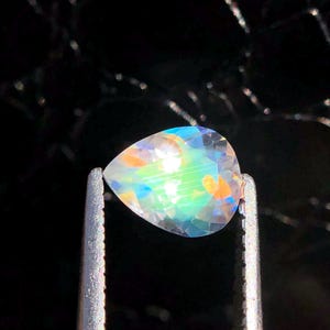 Op de afbeelding: Een peervormige, iriserende edelsteen met een regenboog van kleuren, waaronder blauw, groen en oranje, wordt vastgehouden in een pincet tegen een zwarte achtergrond.