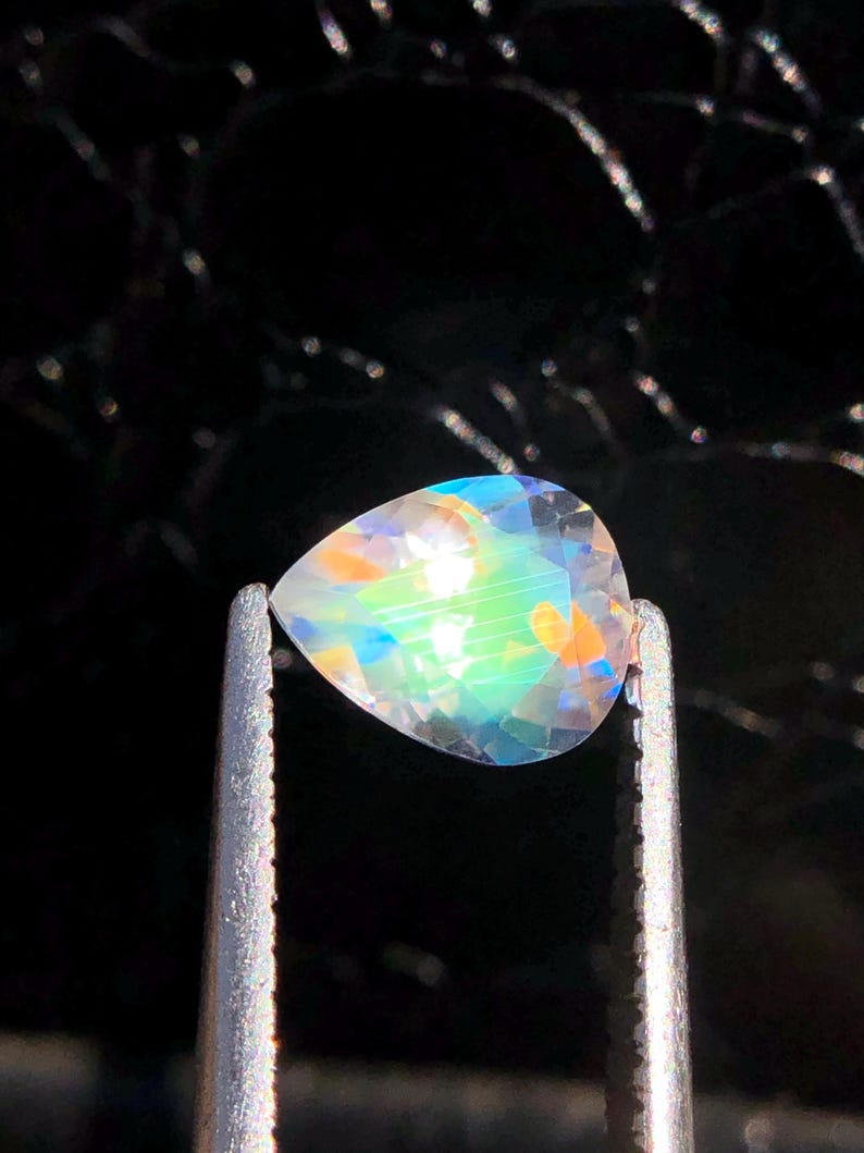 Op de afbeelding: Een peervormige, iriserende edelsteen met een regenboog van kleuren, waaronder blauw, groen en oranje, wordt vastgehouden in een pincet tegen een zwarte achtergrond.