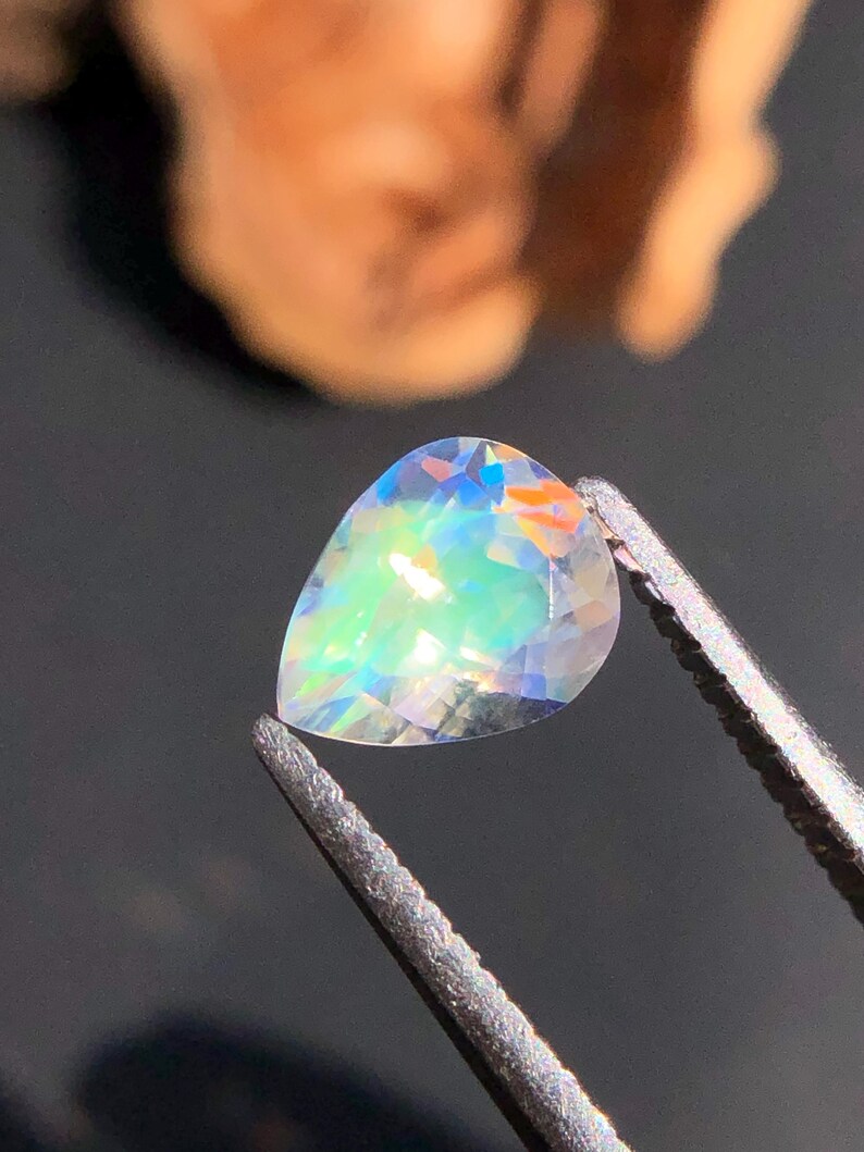Op de afbeelding: Een peervormige, iriserende edelsteen met een regenboog aan kleuren, vastgehouden in een pincet.