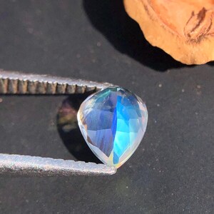 Op de afbeelding: Een regenboogkleurige, traanvormige edelsteen met een blauwe en groene tint. De edelsteen wordt vastgehouden in een pincet tegen een donkere achtergrond.