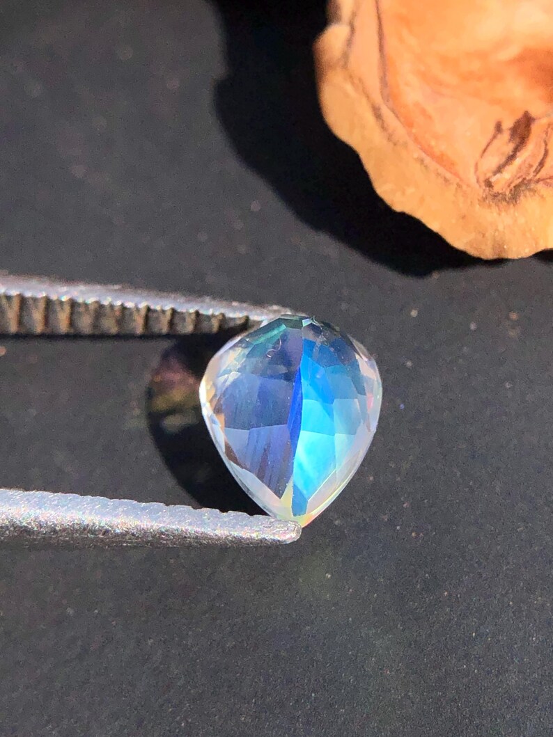 Op de afbeelding: Een regenboogkleurige, traanvormige edelsteen met een blauwe en groene tint. De edelsteen wordt vastgehouden in een pincet tegen een donkere achtergrond.