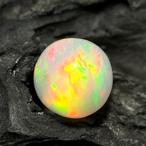 May include: A round, iridescent opal gemstone. The opal displays a play of color with hues of yellow, green, pink, and white. The gemstone is set against a dark, textured background.