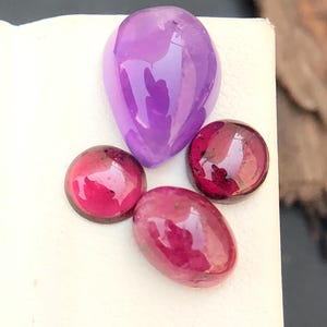 May include: Four polished gemstones: a large, teardrop-shaped amethyst and three smaller, round and oval-shaped garnets. The gems are a deep purple and various shades of red, with a glossy finish. The gems are displayed on a white surface.