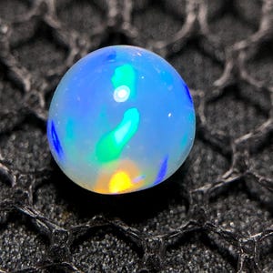 Könnte beinhalten: Ein runder, durchscheinender Cabochon-Edelstein mit einer glatten, polierten Oberfläche. Der Stein zeigt ein Farbspiel mit lebhaften Blitzen in Neonblau, hellem Grün und einem warmen Gelb-Orange vor einem milchig weißen Hintergrund.
