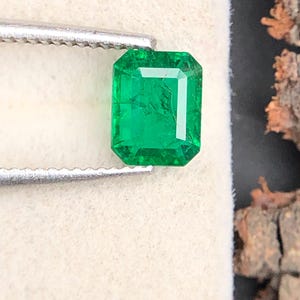 Op de afbeelding: Een heldergroene smaragd, geslepen in een smaragd-vorm, vastgehouden door een zilveren pincet. De edelsteen heeft een rechthoekige vorm met zichtbare facetten. De achtergrond is een zacht, gebroken witte ondergrond met een donker, getextureerd element rechts.