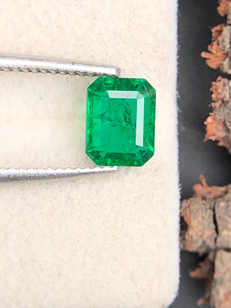 Op de afbeelding: Een heldergroene smaragd, geslepen in een smaragd-vorm, vastgehouden door een zilveren pincet. De edelsteen heeft een rechthoekige vorm met zichtbare facetten. De achtergrond is een zacht, gebroken witte ondergrond met een donker, getextureerd element rechts.