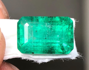 May include: A vibrant, rectangular emerald gemstone with a rich, deep green hue. The gem is held on a white surface, showcasing its clarity and facets. The emerald's cut highlights its brilliance, making it a striking piece of jewelry.