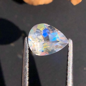 Op de afbeelding: Een regenboogkleurige, peervormige edelsteen met iriseren, vastgehouden in een pincet tegen een zwarte achtergrond.