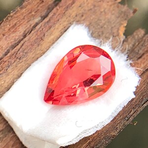 May include: A faceted, teardrop-shaped gemstone in a vibrant orange-red hue. The gem is set on a small, white cloth, resting on a piece of weathered, brown wood. The gem's cut creates multiple reflective surfaces.