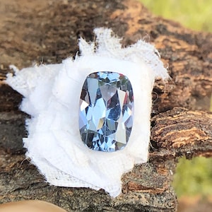 May include: A cushion-cut blue gemstone with a faceted surface. The gemstone is set on a white fabric background.